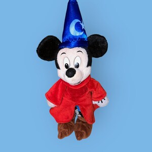 May include: A plush Mickey Mouse toy wearing a blue sorcerer's hat with a crescent moon and star design. He is dressed in a red robe with brown shoes.