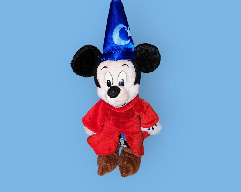 May include: A plush Mickey Mouse toy wearing a blue sorcerer's hat with a crescent moon and star design. He is dressed in a red robe with brown shoes.