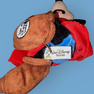 May include: A brown and black plush Mickey Mouse toy wearing a red cape and a brown hat. The toy has a blue tag that says "Walt Disney World."