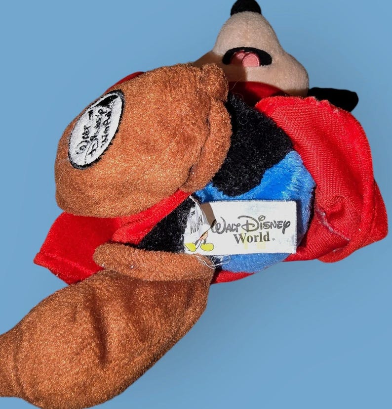 May include: A brown and black plush Mickey Mouse toy wearing a red cape and a brown hat. The toy has a blue tag that says "Walt Disney World."