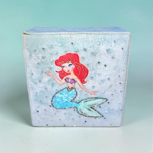 May include: A light blue, square box with a mermaid design. The mermaid has red hair, a blue sequined tail, and a purple top. The box is decorated with silver beads.