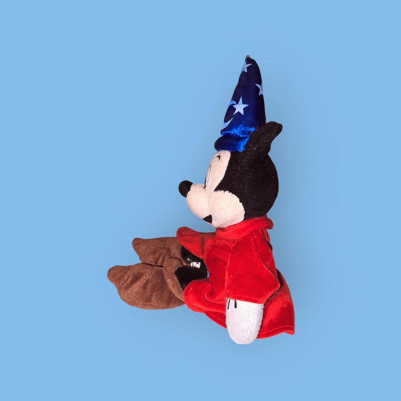 May include: A plush Mickey Mouse toy wearing a blue and white star-patterned wizard hat and a red robe. The toy is sitting with its legs crossed.