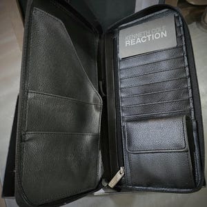 May include: Open black leather wallet with multiple card slots, a zippered compartment, and a small coin pocket. The wallet features the "KENNETH COLE REACTION" logo on a card. The wallet is open, revealing its interior.