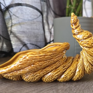 May include: A decorative, ceramic, horn-shaped sculpture in shades of brown and gold. The piece has a textured surface and a glossy finish, with a curled, spiral design. The sculpture is placed on a wooden surface.