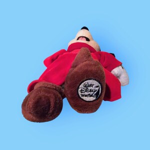 May include: Red plush Mickey Mouse wearing a red robe with a brown face and brown feet. The robe has a "Walt Disney World" logo on the back.