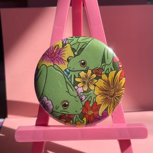May include: A circular badge showing two green frogs amidst vibrant flowers. The badge is set against a pink backdrop and displayed on a pink easel. The flowers include yellow, red, and pink blooms.