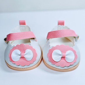 May include: A pair of white shoes with pink scalloped edges and pink bows. The shoes have a white strap with a buckle.