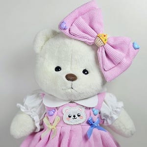 Puede incluir: Un oso de peluche blanco que lleva un vestido rosa con cuello blanco, lazos amarillos y azules y un parche de oso blanco. El oso tiene un lazo rosa con un amuleto de cupcake en la cabeza.