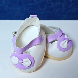 May include: A pair of white doll shoes with purple straps and bows. The shoes have a scalloped edge and a white bow on the front.