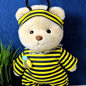 May include: A plush teddy bear wearing a yellow and black striped bee costume with black antennae and yellow pom-poms. The bear has a black headband with a yellow stripe.
