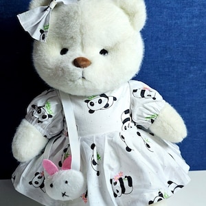 May include: White plush teddy bear wearing a white dress with black and white panda print. The dress has a white bow and a white strap with a small white bunny purse attached.