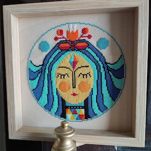 Puede incluir: Un bordado de punto de cruz enmarcado que representa a una mujer estilizada con cabello azul, rostro amarillo y un patrón geométrico colorido en el cuello. La mujer está rodeada de un círculo con un diseño floral en la parte superior.
