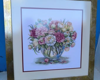 Op de afbeelding: Een ingelijst kruissteekkunstwerk van een bloemstuk in een glazen kom, met roze, witte en gele bloemen. Het kunstwerk is gevat in een goudkleurige lijst met een crèmekleurige passe-partout.