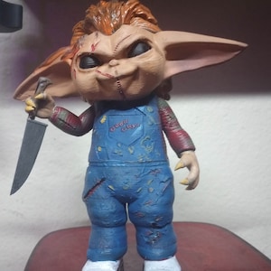 May include: A small, orange-haired, stitched-faced figurine with large ears, holding a knife. The figurine is wearing blue overalls with the words "Good Guy" on the front.