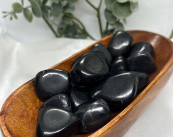 May include: A wooden bowl filled with polished black stones. The stones have a smooth, glossy surface and are oval-shaped. The bowl is made of light brown wood and has a simple, elegant design. Greenery is visible in the background.