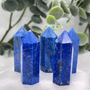 May include: Five polished lapis lazuli crystal towers. The deep blue stones have gold flecks and are cut into hexagonal shapes with pointed tops. The crystals are arranged on a white surface with green foliage in the background.