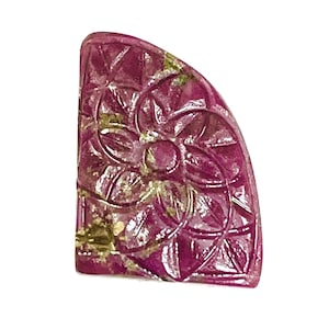 May include: A semi-circular, deep magenta gemstone cabochon featuring a carved, symmetrical floral pattern with intersecting lines. The surface displays natural inclusions and a polished finish, with a flat vertical edge and a curved outer border.