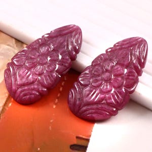 Puede incluir: Dos piedras preciosas en forma de lágrima de color magenta intenso con intrincados patrones florales tallados a mano en sus superficies. Cada piedra muestra un acabado pulido con inclusiones naturales visibles y un diseño de pétalos simétrico.