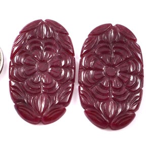 Puede incluir: Dos piedras preciosas ovaladas de color rojo intenso talladas con un patrón floral, colocadas junto a una moneda de cuarto de dólar estadounidense para comparar el tamaño. Las piedras presentan diseños de pétalos simétricos y superpuestos.