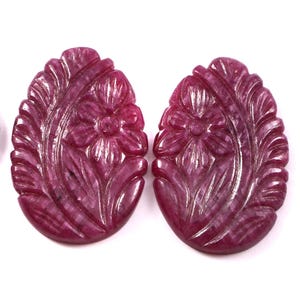 Pode incluir: Duas pedras preciosas esculpidas de cor magenta profunda, de forma oval, com um motivo floral de pétalas e folhas. Uma moeda americana de um quarto de dólar está posicionada à esquerda para escala, medindo 2,43 cm de diâmetro.