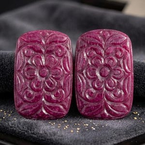 Puede incluir: Dos piedras preciosas rectangulares de color magenta intenso con intrincados patrones florales tallados a mano. Cada piedra muestra un diseño simétrico con una flor central rodeada de hojas estilizadas y líneas curvas con acabado pulido.