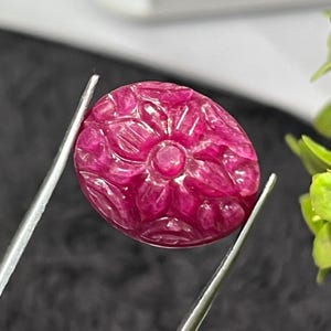 Puede incluir: Una piedra preciosa ovalada de color magenta intenso con una intrincada talla floral en su superficie. La piedra muestra una calidad translúcida con inclusiones naturales visibles y está sujeta por unas pinzas de metal plateado.