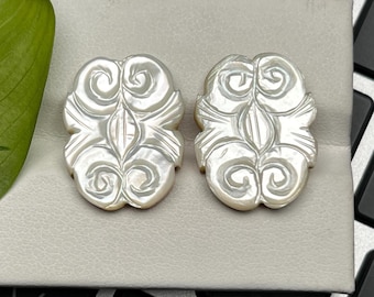 May include: Pair of iridescent, pearly white stud earrings. Each earring features an ornate, symmetrical design with swirling patterns and a central, elongated shape. The earrings are displayed on a light gray surface.