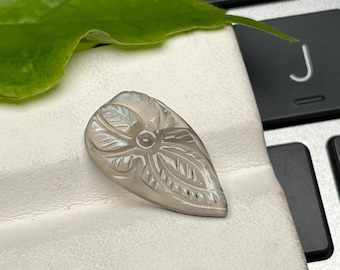 May include: A carved, teardrop-shaped gemstone with a floral and leaf design. The stone is a pale, translucent gray color and rests on a white surface. A green leaf is visible in the background, along with a keyboard.