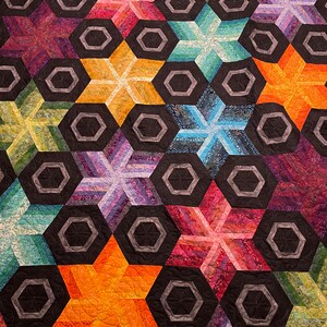 Puede incluir: Una colcha con un patrón repetitivo de estrellas y hexágonos coloridos. Las estrellas están hechas de diferentes telas y tienen un efecto 3D. Los hexágonos son negros con un contorno gris.