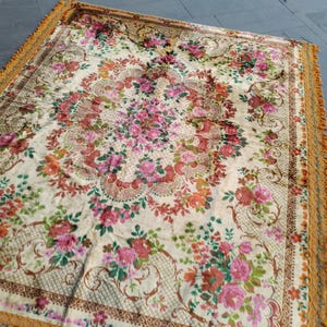 Puede incluir: Una alfombra rectangular con un estampado floral en tonos rosa, rojo y naranja sobre un fondo crema. La alfombra tiene un borde decorativo y un fleco de color amarillo dorado. La alfombra está colocada sobre una superficie gris.