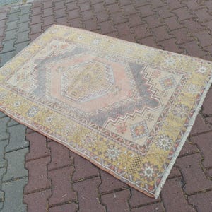 Op de afbeelding: Rechthoekig vintage tapijt met een centraal geometrisch ontwerp in perzik en grijs, omgeven door een gele rand met bloemen- en geometrische patronen. Het tapijt heeft een versleten uitstraling, met tekenen van slijtage.