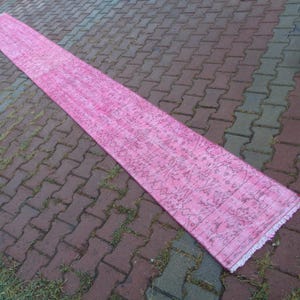 May include: A long pink runner rug with a floral pattern and fringed edges. The rug is laid out on a brick patio.