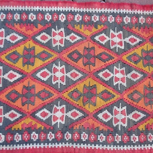 K&ouml;nnte beinhalten: Ein bunter Teppich mit einem geometrischen Muster in Rot-, Orange-, Gelb-, Wei&szlig;- und Schwarzt&ouml;nen. Der Teppich hat ein rautenf&ouml;rmiges Design mit komplizierten Details.