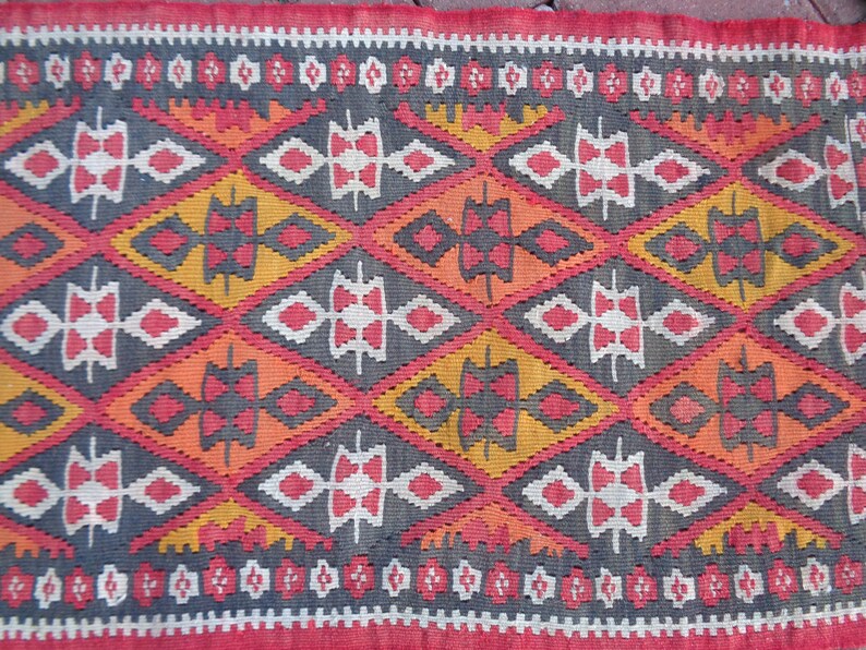 K&ouml;nnte beinhalten: Ein bunter Teppich mit einem geometrischen Muster in Rot-, Orange-, Gelb-, Wei&szlig;- und Schwarzt&ouml;nen. Der Teppich hat ein rautenf&ouml;rmiges Design mit komplizierten Details.