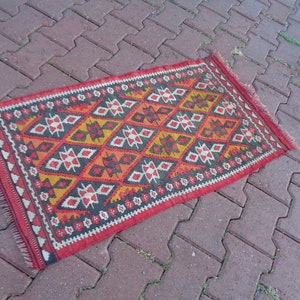K&ouml;nnte beinhalten: Ein roter, orangefarbener, gelber, wei&szlig;er und schwarzer Teppich mit einem Fransenrand. Der Teppich liegt auf einer Terrasse aus Ziegeln.