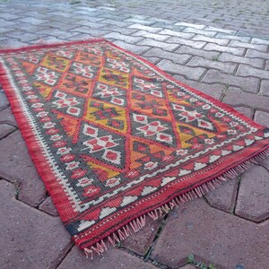 K&ouml;nnte beinhalten: Ein roter, orangefarbener, gelber und schwarzer Teppich mit einem Fransenrand. Der Teppich liegt auf einem Ziegelsteinweg.