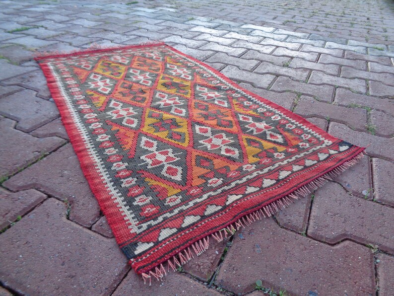 K&ouml;nnte beinhalten: Ein roter, orangefarbener, gelber und schwarzer Teppich mit einem Fransenrand. Der Teppich liegt auf einem Ziegelsteinweg.