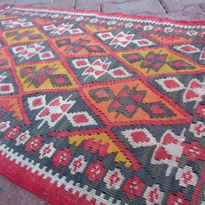 K&ouml;nnte beinhalten: Ein roter, orangefarbener, wei&szlig;er und grauer Teppich mit geometrischem Muster. Der Teppich ist in traditioneller Weise gewebt und hat einen Fransenrand.