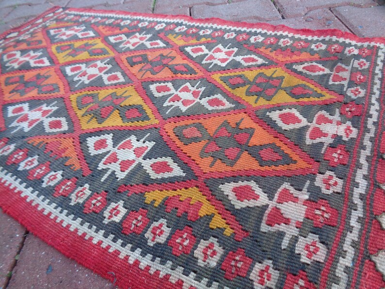 K&ouml;nnte beinhalten: Ein roter, orangefarbener, wei&szlig;er und grauer Teppich mit geometrischem Muster. Der Teppich ist in traditioneller Weise gewebt und hat einen Fransenrand.