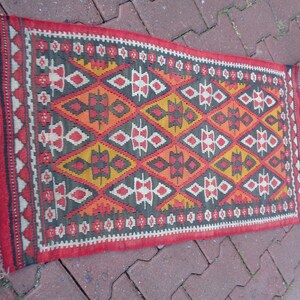 K&ouml;nnte beinhalten: Ein roter, orangefarbener, gelber und schwarzer Teppich mit geometrischem Muster. Der Teppich hat einen Fransenrand.