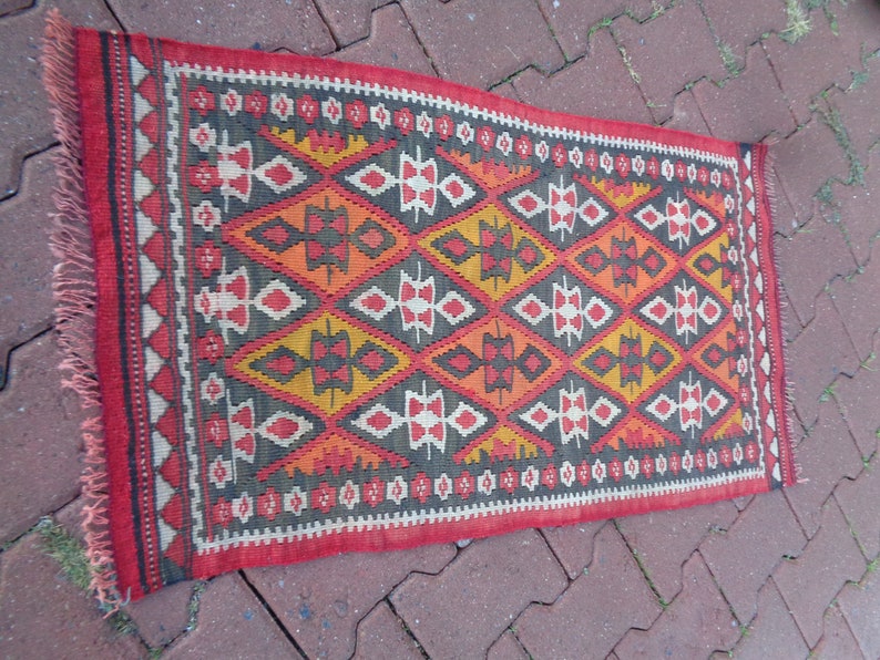 K&ouml;nnte beinhalten: Ein roter, orangefarbener, gelber und schwarzer Teppich mit geometrischem Muster. Der Teppich hat einen Fransenrand.