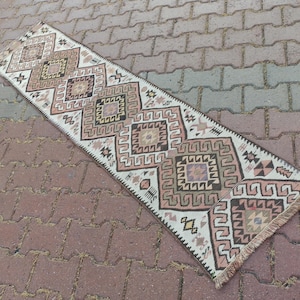 May include: A rectangular, patterned rug with a cream-colored background and geometric designs in shades of brown, pink, and purple. The rug has a fringed edge and is placed on a brick surface.