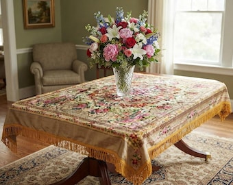 Könnte beinhalten: Eine rechteckige Tischdecke mit beigem Untergrund, einem mehrfarbigen Blumenmuster und goldfarbenen Fransen an den Rändern. In der Mitte steht eine klare Vase aus geschliffenem Glas mit einem großen Strauß aus rosa, roten, weißen und blauen Blumen.