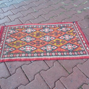 K&ouml;nnte beinhalten: Ein bunter Teppich mit einem geometrischen Muster in Rot-, Orange-, Gelb-, Schwarz- und Wei&szlig;t&ouml;nen. Der Teppich hat einen Fransenrand und liegt auf einer Ziegelsteinfl&auml;che.