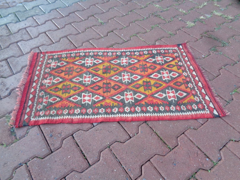 K&ouml;nnte beinhalten: Ein bunter Teppich mit einem geometrischen Muster in Rot-, Orange-, Gelb-, Schwarz- und Wei&szlig;t&ouml;nen. Der Teppich hat einen Fransenrand und liegt auf einer Ziegelsteinfl&auml;che.