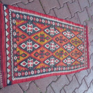 K&ouml;nnte beinhalten: Ein traditioneller t&uuml;rkischer Kilim-Teppich mit einem geometrischen Muster in Rot-, Orange-, Gelb-, Schwarz- und Wei&szlig;t&ouml;nen. Der Teppich hat einen Fransenrand.