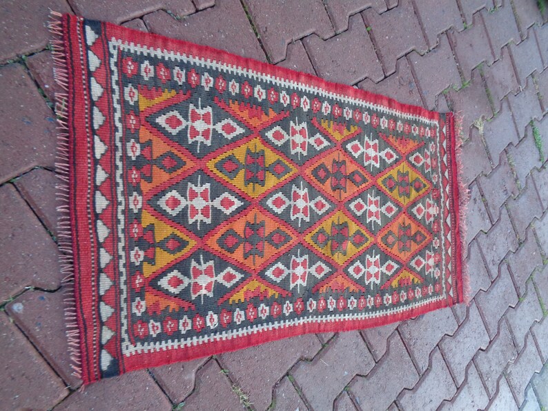 K&ouml;nnte beinhalten: Ein traditioneller t&uuml;rkischer Kilim-Teppich mit einem geometrischen Muster in Rot-, Orange-, Gelb-, Schwarz- und Wei&szlig;t&ouml;nen. Der Teppich hat einen Fransenrand.