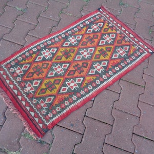 K&ouml;nnte beinhalten: Ein roter, orangefarbener, gelber und schwarzer Teppich mit Fransen an den R&auml;ndern. Der Teppich hat ein geometrisches Muster mit Diamantenformen und floralen Motiven.