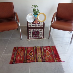 May include: Two brown leather chairs with chrome legs, a small table with a brown and white patterned runner, and two white plates with a gold rim and a small decorative figurine on top of the table. A red, orange, yellow, and black patterned runner is on the floor.