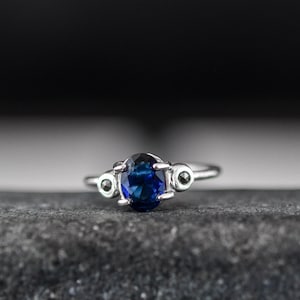 Op de afbeelding: Een zilveren ring met een grote blauwe ovale edelsteen en twee kleinere zwarte edelstenen aan weerszijden.